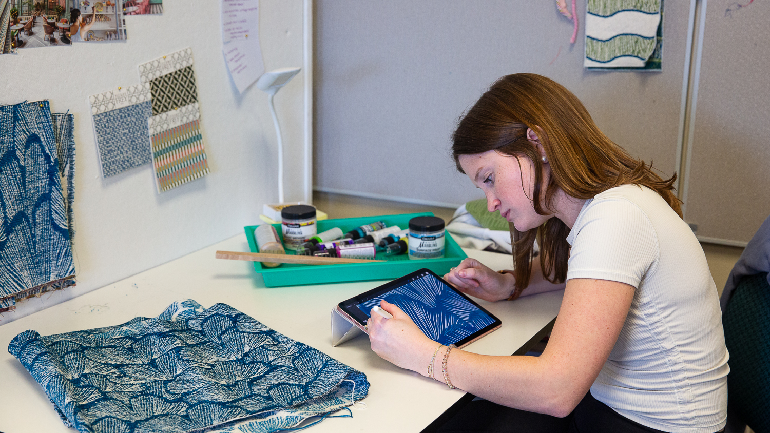 Tessa Dionne uses her tablet to update the design on the fabric beside her on the table, which is a deep blue with white stitching to create a leaf-like pattern.