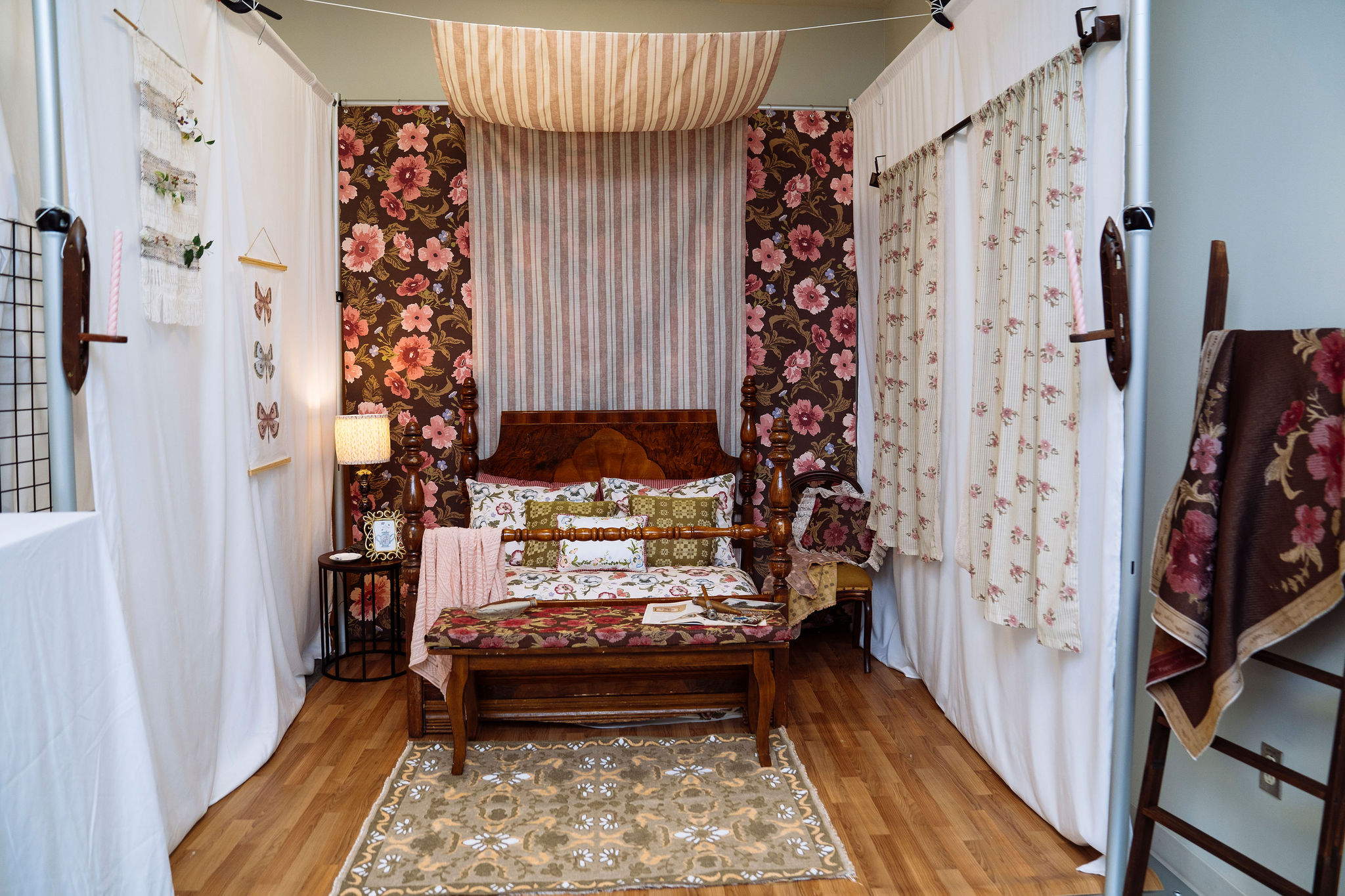 A booth is styled like a cozy bedroom with floral-patterned bedding, pillows, and curtains, a wooden bed, side tables with lamps, a beige patterned rug, and wood flooring. The room features warm, earthy tones and layered textiles.