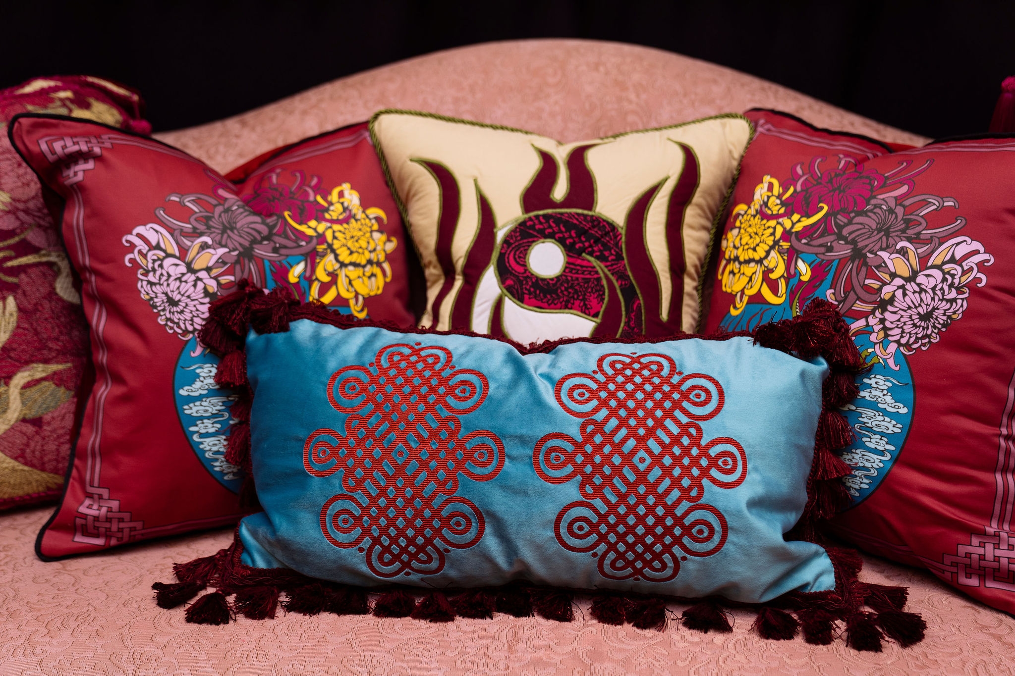 A blue pillow with red fringe and stitching is surrounded by other decorative pillows on a pink lounge chair.