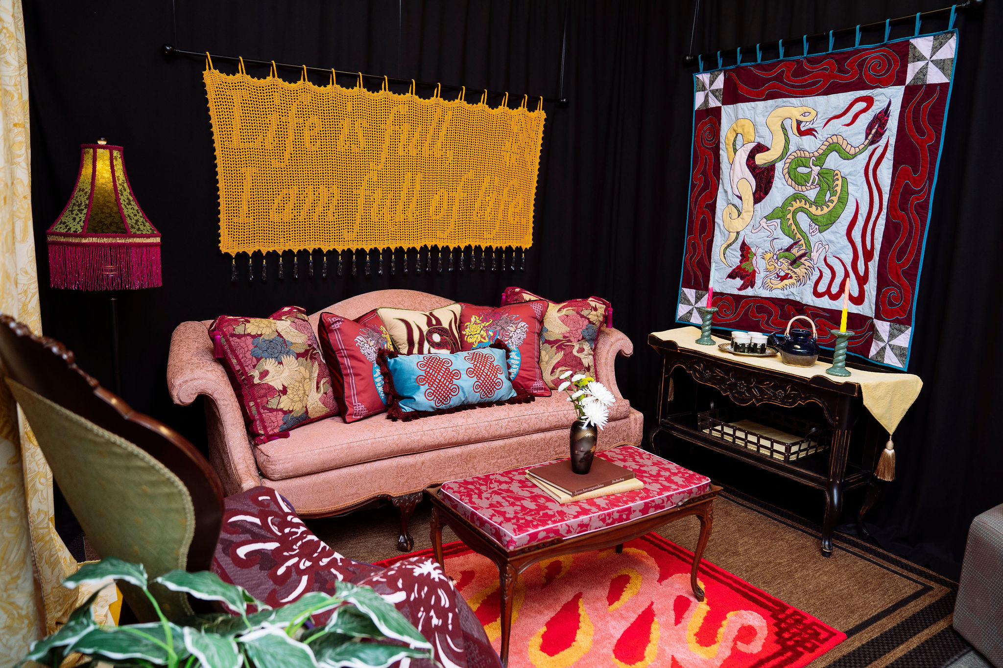 A room with a pink couch and coffee table, with a yellow crocheted art piece that reads "Life is full, I am full of life." The couch is decorated with red and blue patterned pillows. A green lampshade with pink fringe is on the left f the couch. To the right is a quilted tapestry depicting a dragon and a snake hung above a side table.
