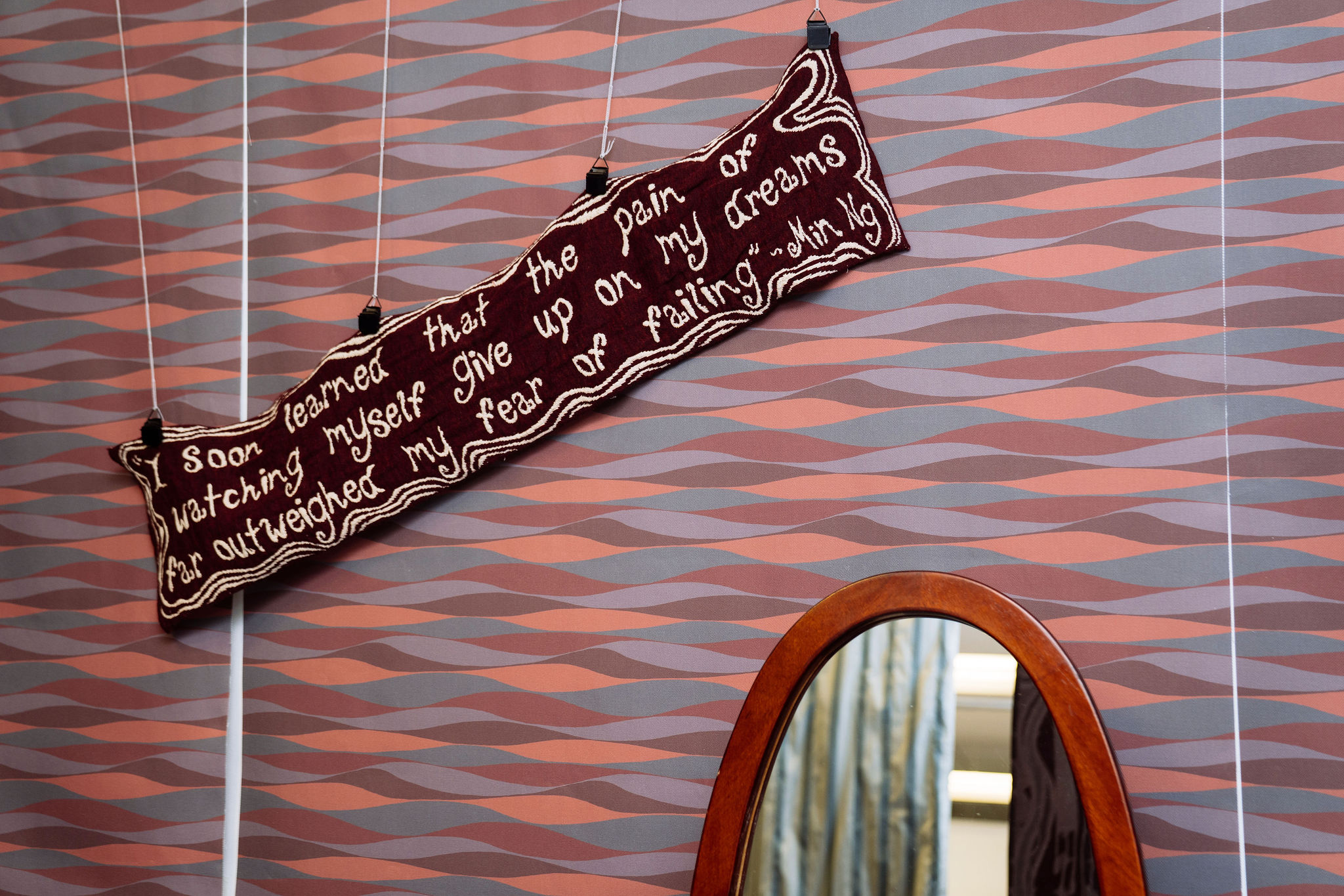 A maroon woven pillow hangs on patterned wallpaper above a wooden-framed mirror. The banner reads: "Soon learned that the pain of watching myself give up on my dreams far outweighed my fear of failing."