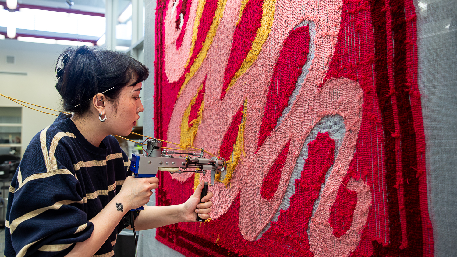 A student works on a red rug with twirling pink lines with a tufting gun.