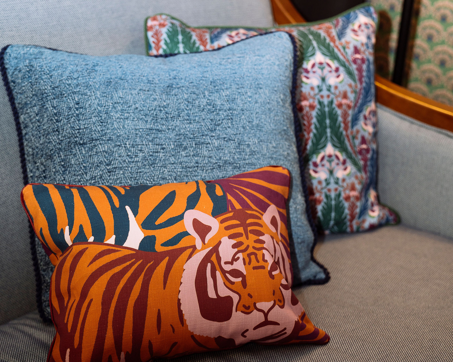 A decorative sofa features three cushions: one with a colorful tiger illustration, one with a blue textured pattern, and one with a floral pattern in the background.