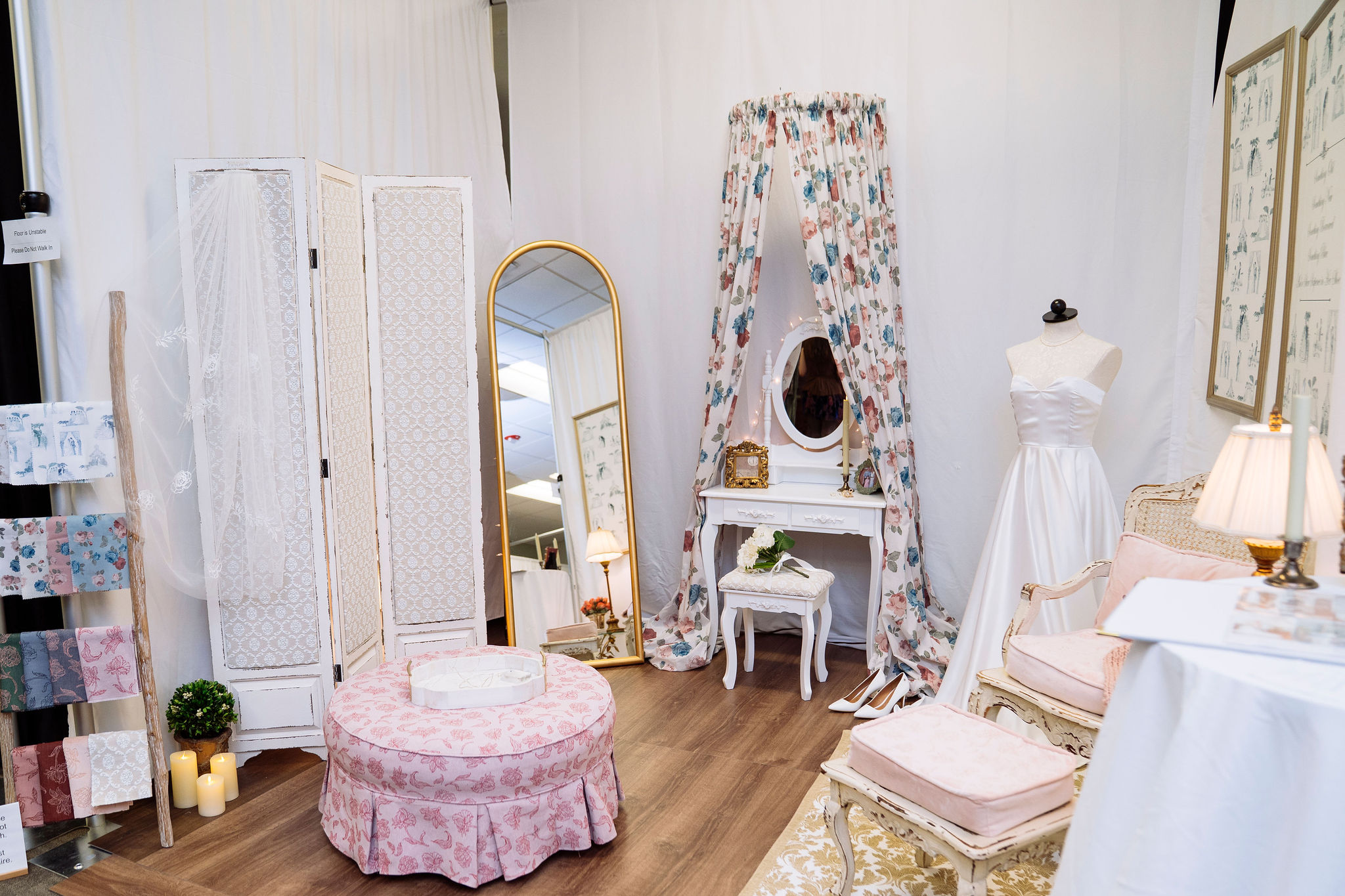 A booth styled like a bridal dressing room with a white gown on a dress form, a vanity table with a floral canopy, a tall mirror, a pink ottoman, a folding screen and elegant armchairs.