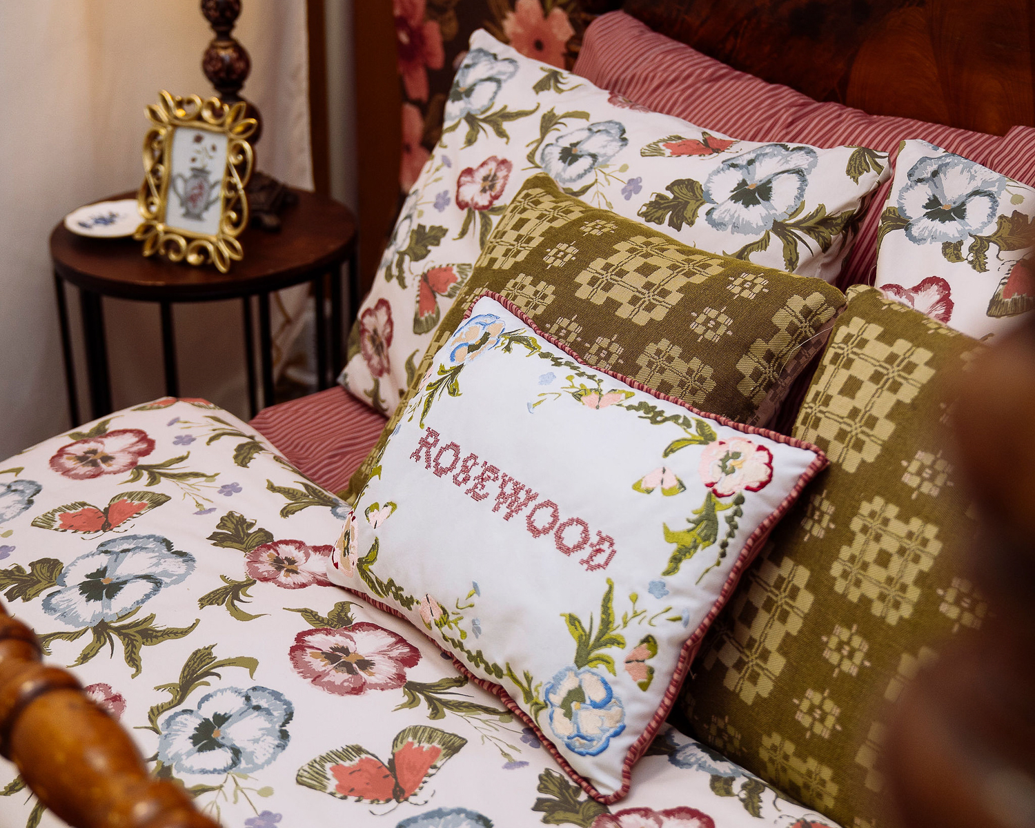 A close up of green, pink and white decorative pillows on a bed. The front-facing pillow has the word "Rosewood" stitched in red and surrounded by florals. In the background, there is a dark brown side table with a golden frame atop it.