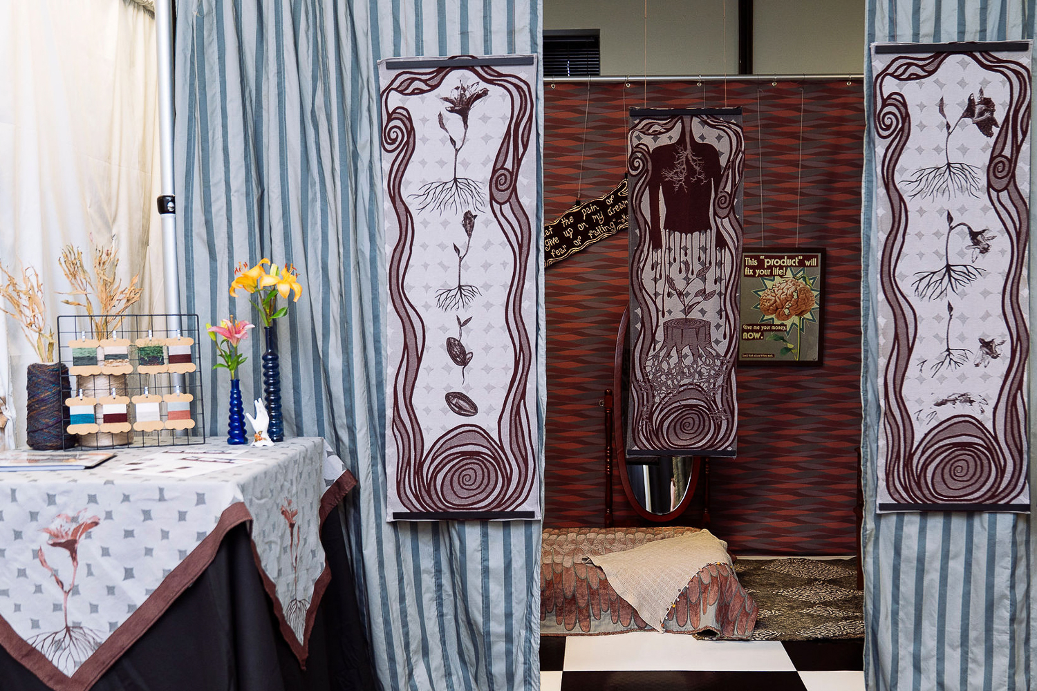 A booth decorated with patterned fabric panels, a small bed, a table with folded textiles, vases with flowers and wall hangings. The setting has earthy tones.