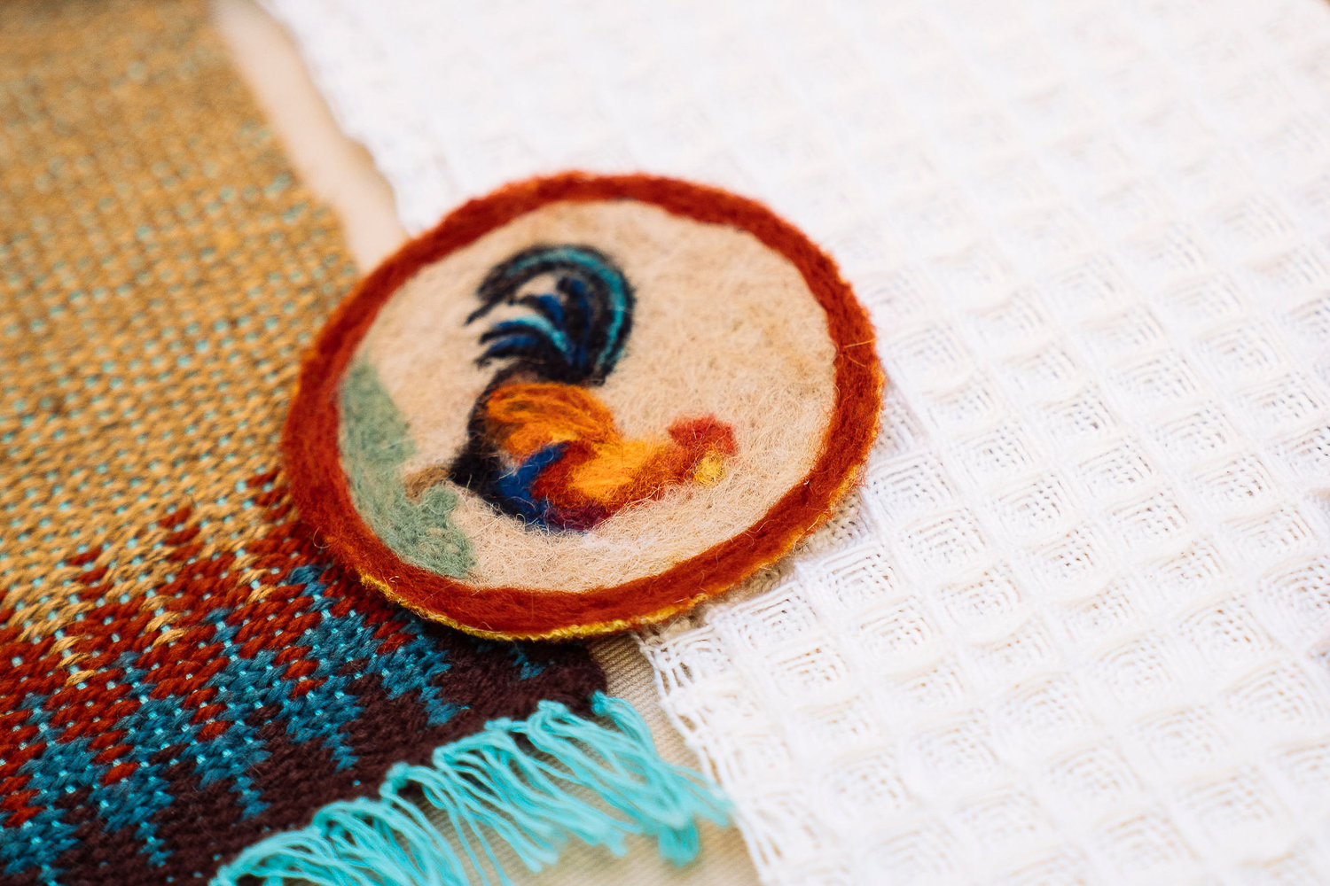 A round felted coaster with a rooster pattern bordered in red, sits on top of a white textured fabric next to a multicolored woven textile with blue, brown, and beige threads.