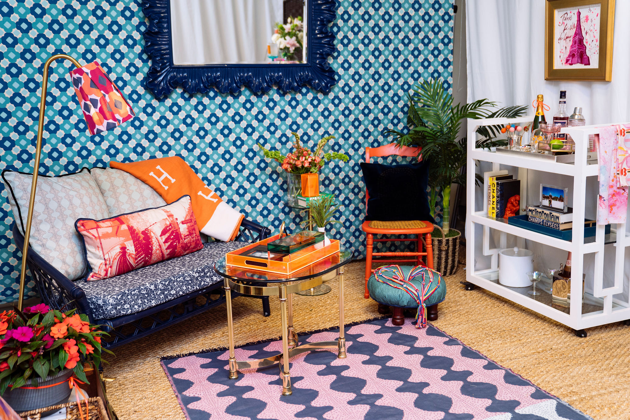 A colorful, eclectic living room with a blue patterned wall, small sofa with patterned cushions, orange blanket, bright tray table, pink and blue rug, bar cart, plants and various decorative accents.