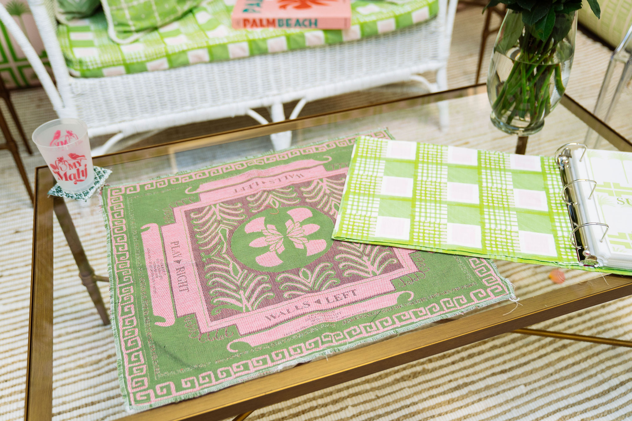 A glass-top table with a pink and green Palm Beach woven game board, a binder with green and and a cup. A white wicker sofa with patterned cushions is in the background.