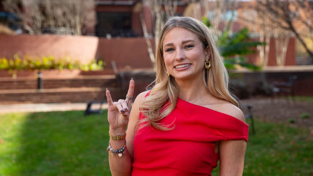 Sutton Hodges smiles while wearing a red dress standing outdoors on a grassy area, making the "Wolfpack" hand sign. She wears gold jewelry and her long blonde hair is down. Trees and a brick building are in the background.
