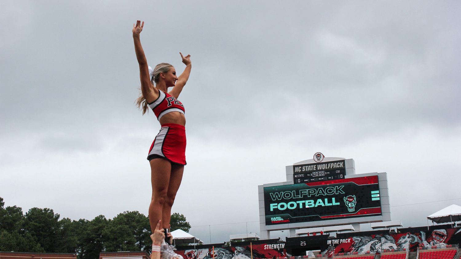 Sutton Hodges wears a red and black uniform while performing a cheerleading stunt on a football field. The scoreboard behind her reads "NC State Wolfpack Football." Trees and cloudy skies are visible in the background.