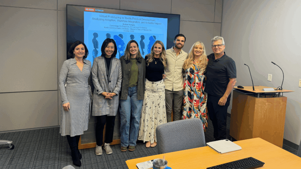 Sutton Hodges and six other people stand together and smile in a conference room in front of a presentation screen displaying a slide about Hodges' Master's Thesis on virtual participation in new product development.
