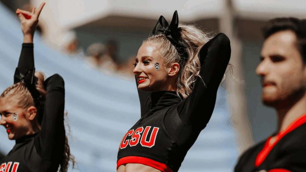 Sutton Hodges wears a black and red NCSU uniform and smiles with her arms raised, standing among fellow cheerleaders during a sports event. She has a large black bow in her hair and face stickers with a Wolfpack logo.