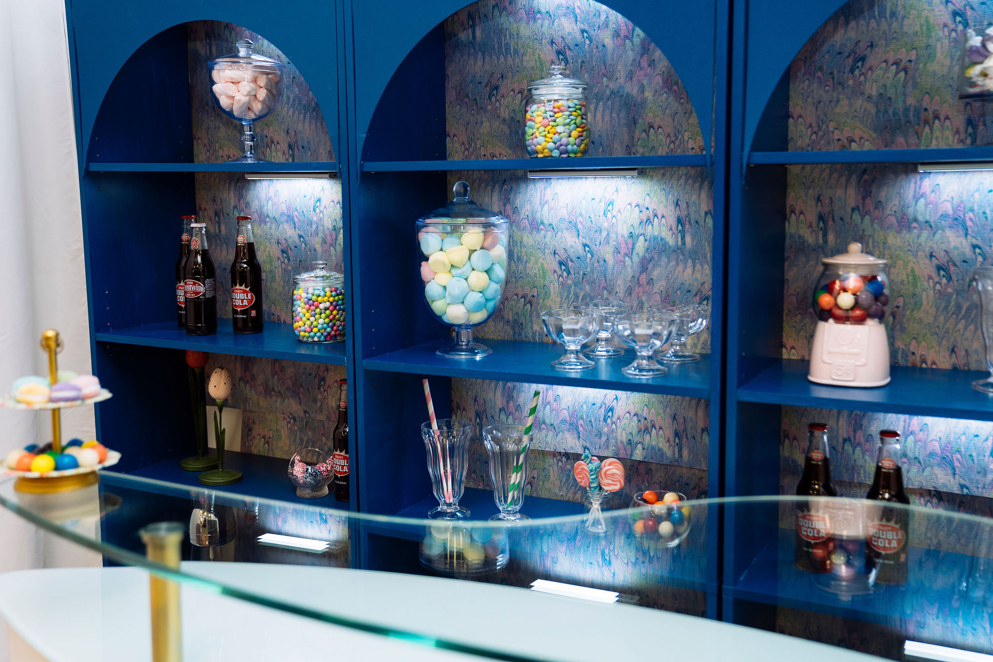 A blue shelving unit displays colorful candies in glass jars, bottles of soda, empty glasses and lollipops, behind a glass counter in a retro-style candy shop. Colorful wallpaper is on the back wall of the shelves.