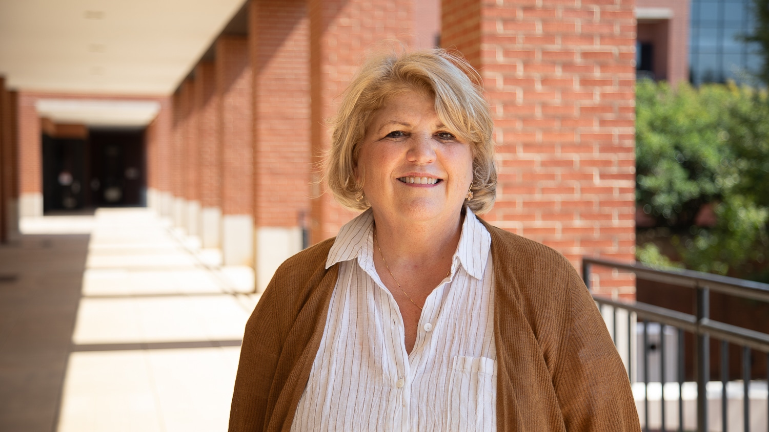 Karen Leonas stands outside in a sunlit breezeway, wearing an off-white blouse and brown sweater, with brick columns and greenery in the background.