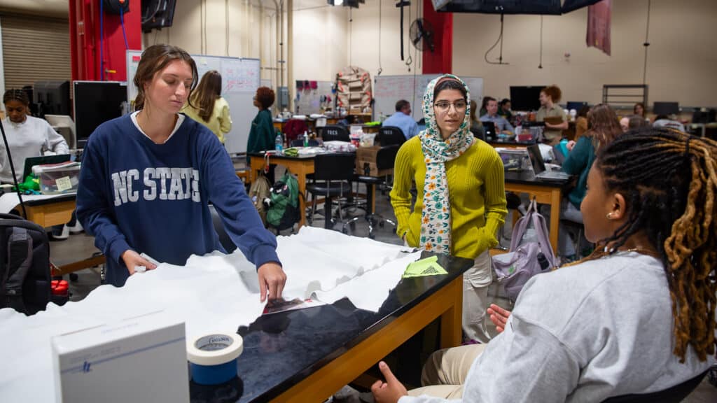 Devon Poulin, Fatemeh Heydari and Jordan Everett work together at a table covered in white knit fabric in the Senior Design Lab. Poulin adjusts the fabric. The room is filled with equipment, supplies and other students in the background.