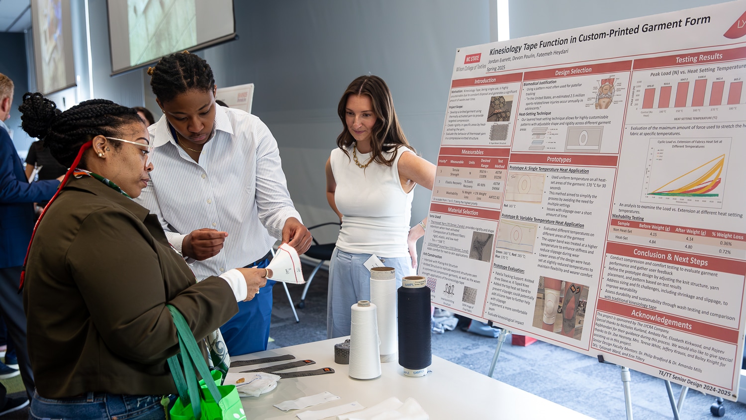 Jordan Everett shows a knit sample to a person. Devon Poulin stands to her right. The poster visible behind them is titled 