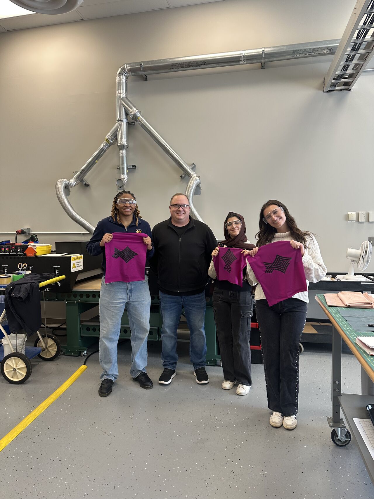 Four people stand and smile in a lab. Devon Poulin, Jordan Everett and Fatemeh Heydari hold up magenta textile swatches with a black geometric design. They all wear protective glasses.