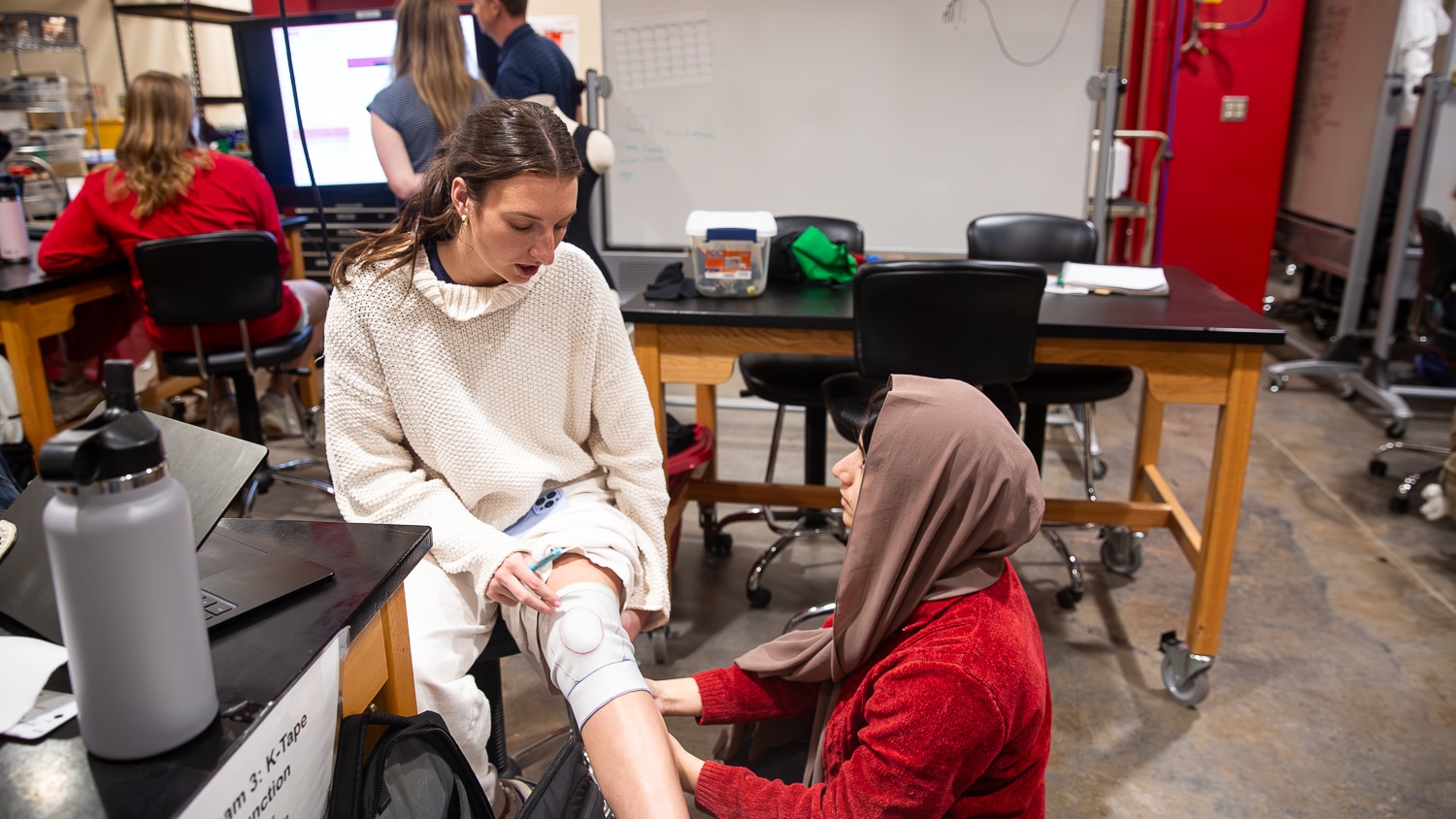 Devon Poulin is seated in the Senior Design Lab while Fatemeh Heydari kneels and holds a knit prototype with red and blue markings to her knee. The room has lab tables, laptops, and other people working in the background.