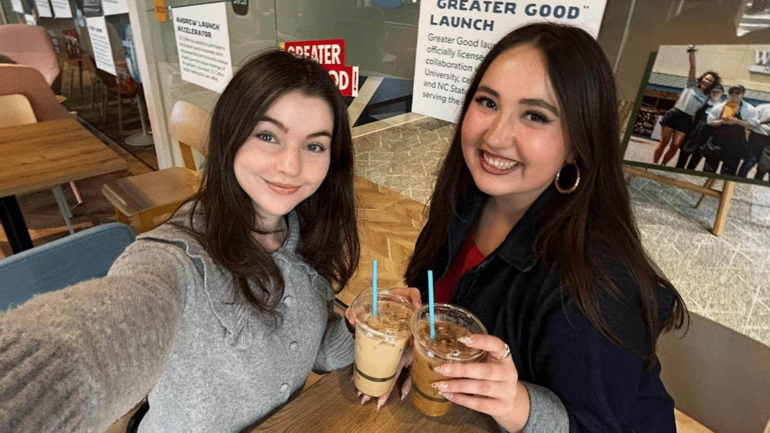 Two students sit at a wooden table at 321 Coffee on Centennial Campus. They are each holding an iced coffee drink and smiling at the camera for a selfie. On the left is a student with long brown hair wearing a grey knitted sweater. On the right is a student with long dark hair, hoop earrings, and a black jacket.