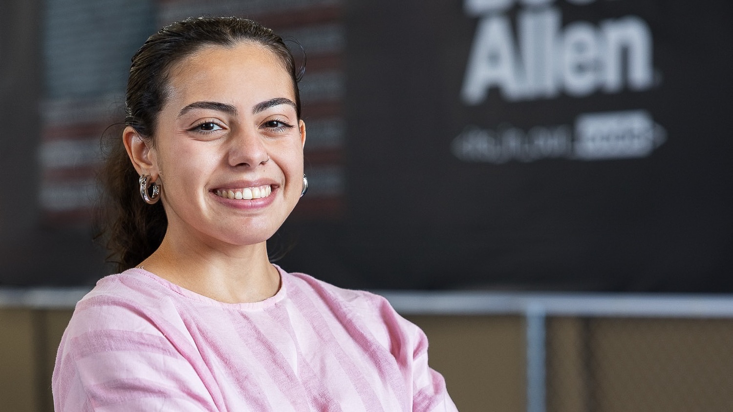 Nicole Rosario-Ortiz wears her hair pulled back, wearing a pink striped shirt and hoop earrings, smiles confidently with arms crossed. The background is blurred with "Allen" visible on the wall.