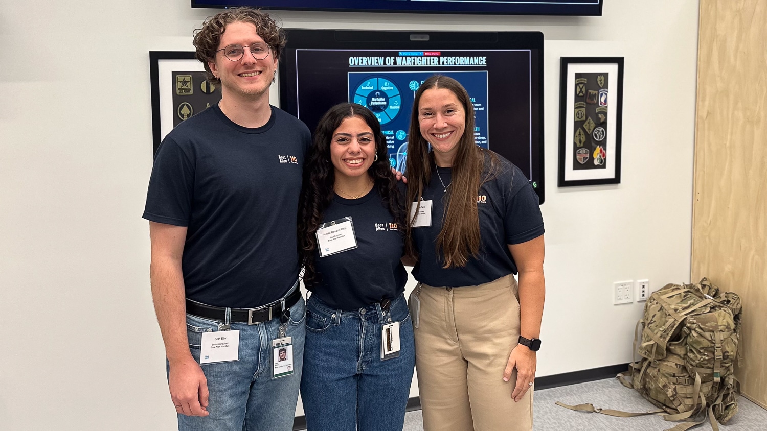 Nicole Rosario-Ortiz and two other people, all wearing navy blue Booz-Allen t-shirts, stand together indoors. Behind them is a large screen displaying with infographics titled "Overview of Warfighter Performance." A camouflage backpack sits on the floor nearby.
