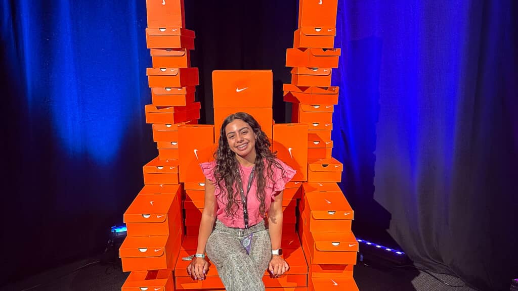 Nicole Rosario-Ortiz sits on a thrown made out of Nike shoeboxes and smiles at the camera. She wears a Nike lanyard. A black backdrop with blue light is visible behind her.