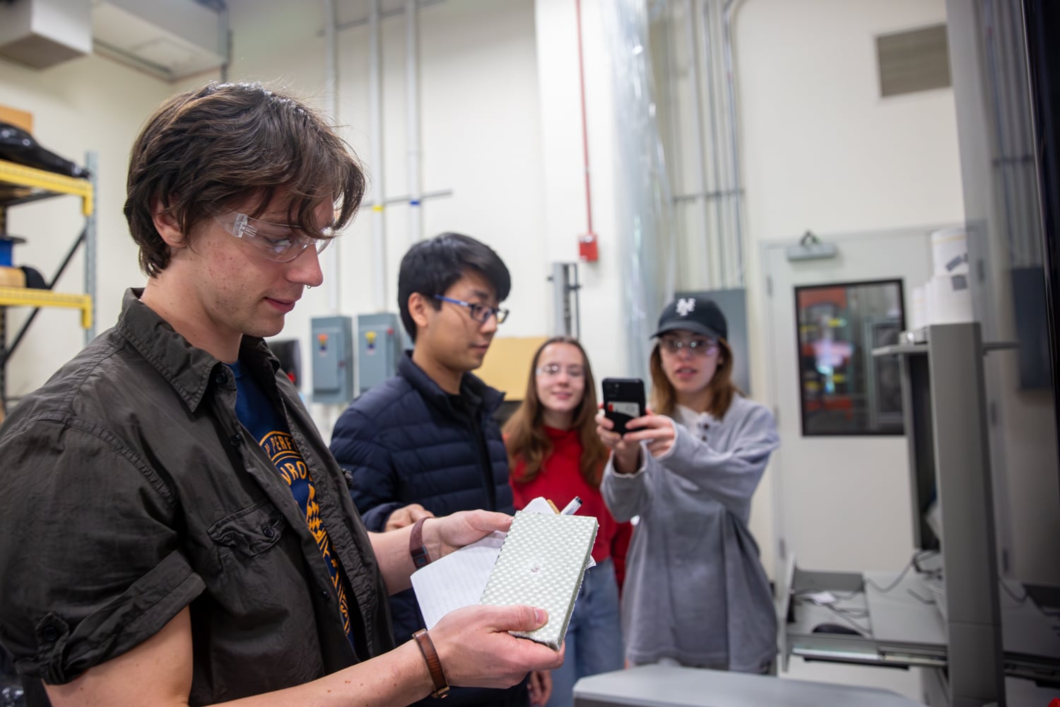 Four people in a lab; one is holding a sample that has just been tested and a notebook, another is looking on, while two others in the background smile and take a photo with a phone. All are wearing glasses or protective glasses.