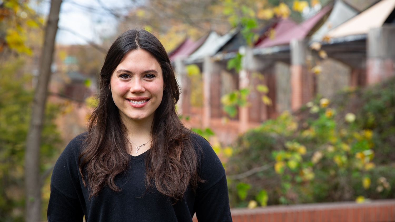 Annie Hoyt wears a black top and smiles while standing outdoors in front of greenery and a row of brick pillars with awnings.