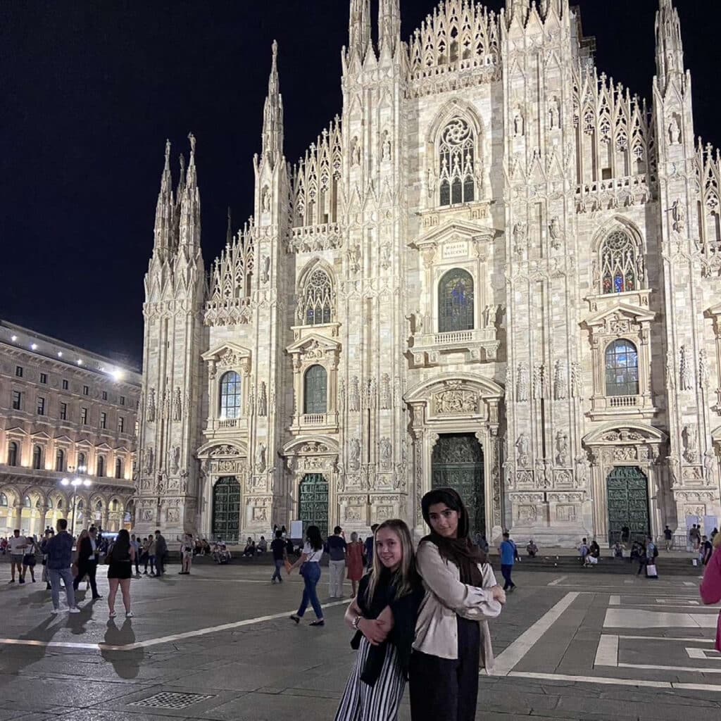 Fatemeh Heydari and another person stand back-to-back with arms crossed at night in front of the illuminated, ornate facade of Milan Cathedral (Duomo di Milano) in Italy. Several people are walking around the square.