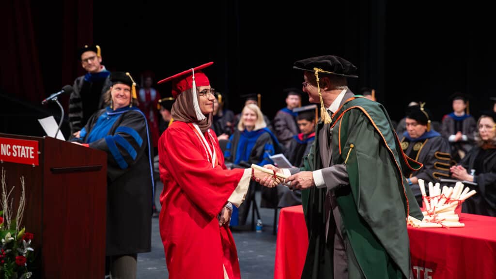 Fatemeh Heydari wears a red cap and gown and shakes hands with David Hinks on stage during a commencement ceremony. Other faculty and rolled facsimile diplomas are visible in the background.