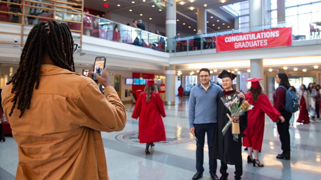 A person takes a photo of a graduate in a cap and gown holding flowers, standing with another person in Talley Student Union with "Congratulations Graduates" banner. Other graduates are visible in the background.