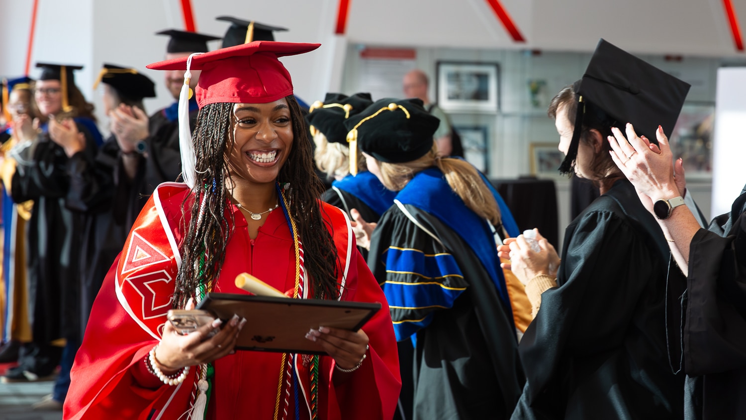 Naomi Southerland wears red cap and gown and holds a rolled facsimile diploma and a frame and walks through a "tunnel" of applauding faculty and staff members dressed in academic regalia during a commencement ceremony.