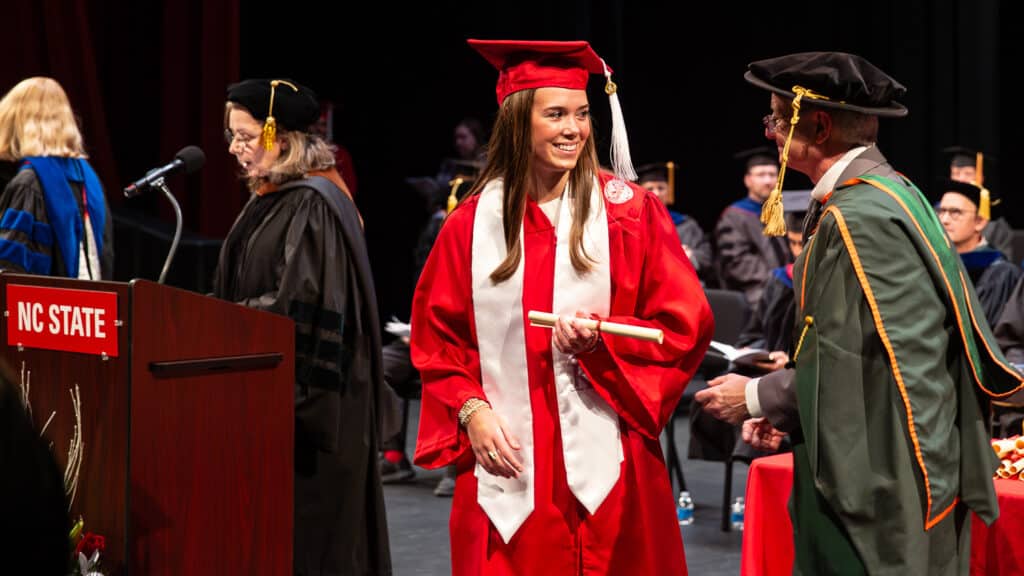 A smiling graduate in a red cap and gown receives their diploma on stage at a Wilson College of Textiles commencement ceremony, surrounded by faculty in academic regalia.