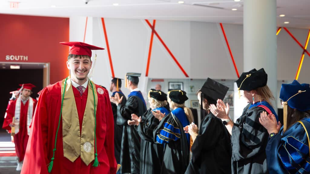 Nate Wolfe wears a red cap and gown and smiles while walking past faculty members in academic regalia who are clapping in a brightly lit hallway.