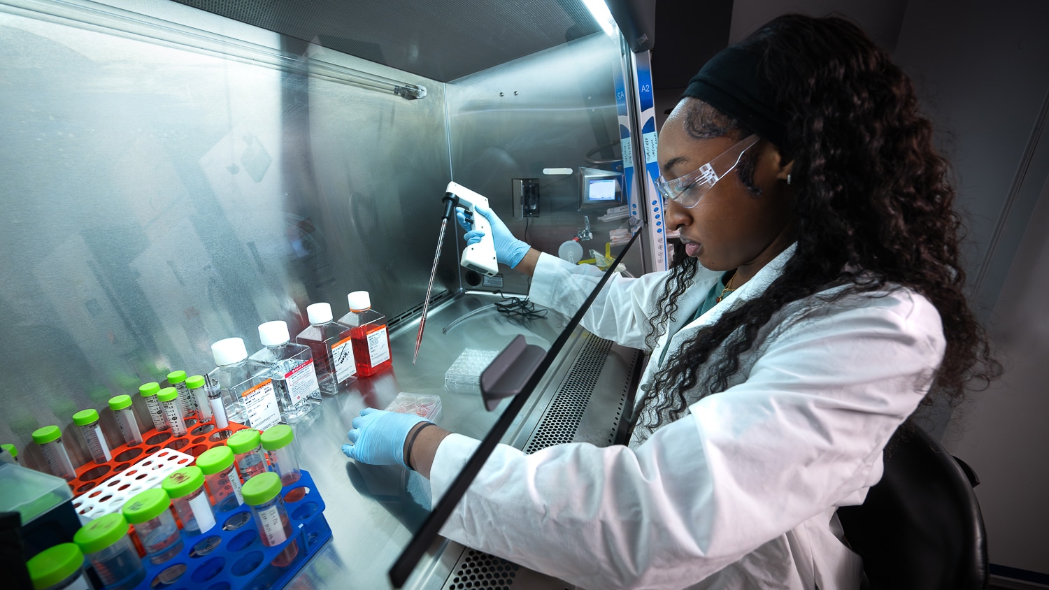 A person wearing a lab coat, gloves and safety goggles uses a pipette in a laboratory, working with samples and test tubes under a fume hood with various containers and scientific equipment.