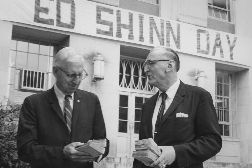William Edward Shinn (left) and Joe Porter (right) stand outside a building, each holding a stack of papers. A large banner reading "ED SHINN DAY" hangs above the entrance to the building behind them. Both men are wearing suits and glasses in the historic, black and white photo.