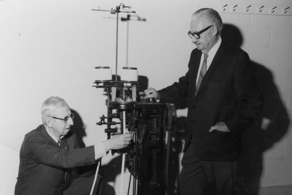 In a historic, black and white photo, William Edward Shinn and Malcom E. Campbell examine and operate a large mechanical knitting device in a room with a plain wall. One man is kneeling and adjusting the machine, while the other stands on the right side of it, watching. Both men wear suits.