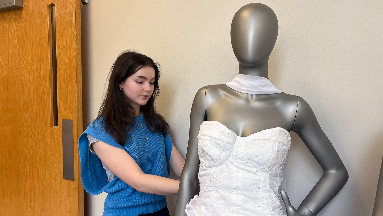 Maylan Maxwell, wearing a blue, knitted sleeveless top, adjusts the white, sleeveless garments on a silver mannequin. Behind her is a white wall and wooden door.