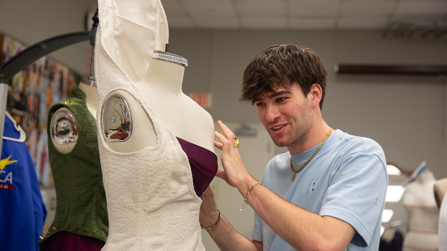 Sam Koziarz-Catz smiles as he adjusts a purple dress on a mannequin in a fashion studio in the Wilson College of Textiles, surrounded by other clothing and dress forms.