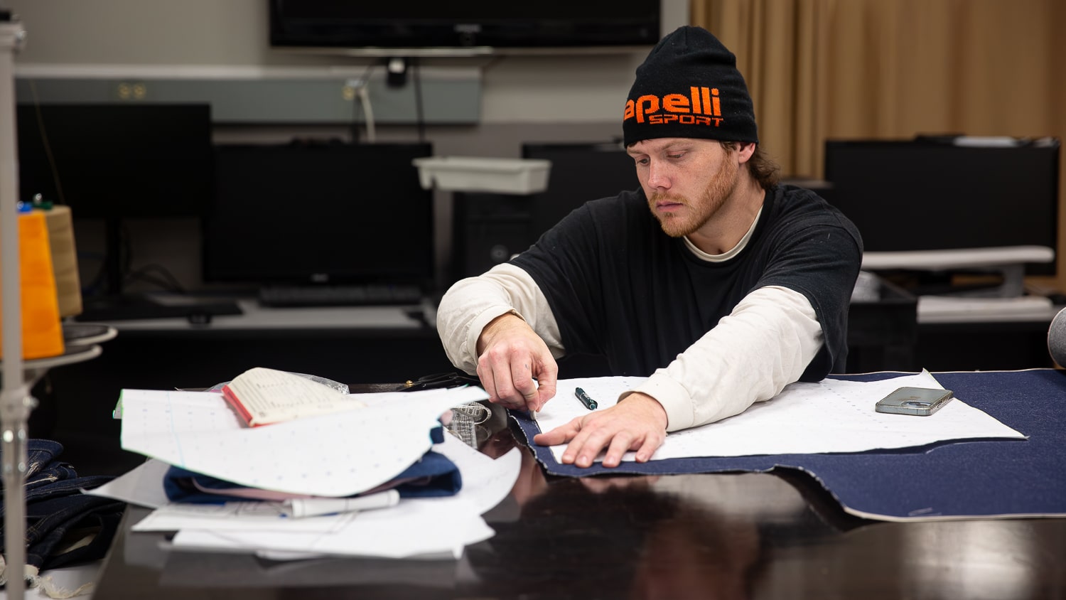 Kaleb Spivey draws a line with marking chalk on fabric at a workstation in a fashion studio, with papers, a notebook and tools scattered on the table. Computer monitors are visible in the background.