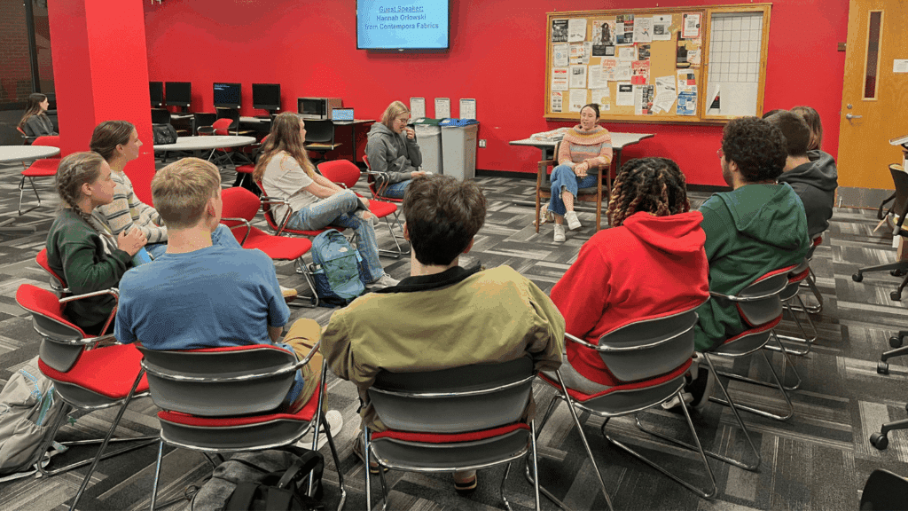 Students sit in chairs in a semicircle, all facing a young woman wearing a pastel sweater. The students, wearing casual clothing like jeans and sweatshirts, are in a red room with computer and bulletins along the walls.