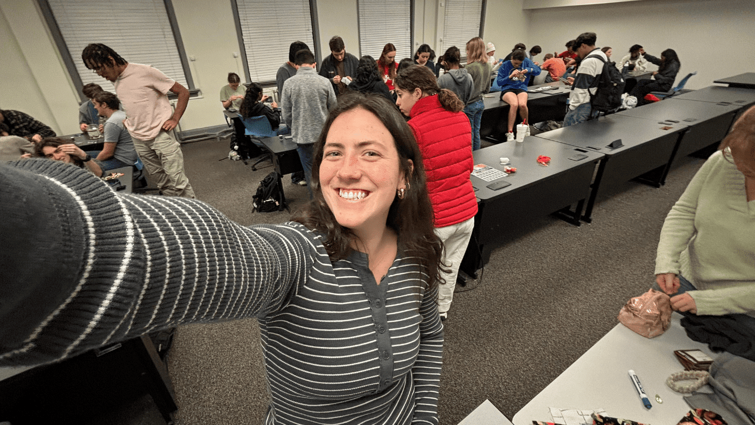 Penny Kudlak smiles as she takes a selfie during a meeting of students in a large, white and grey room. She is wearing a grey shirt with white stripes. Students work on sewing projects at two long tables behind her.
