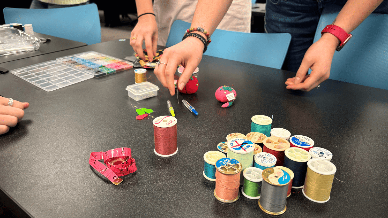 A black table scattered with sewing and mending supplied. A pair of hands reaches for a spool of thread among a collection of colorful spools. There are a few pincushions and a bead organizer on the table, which is bordered with blue chairs.
