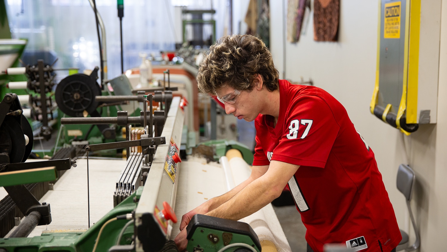 Will Swanson leans down to adjust a loom in the Weaving Lab. He wears a red NC State jersey and the lab stretches out behind him with various machines and equipment. His profile is shown as he works with a loom in the middle of the frame.