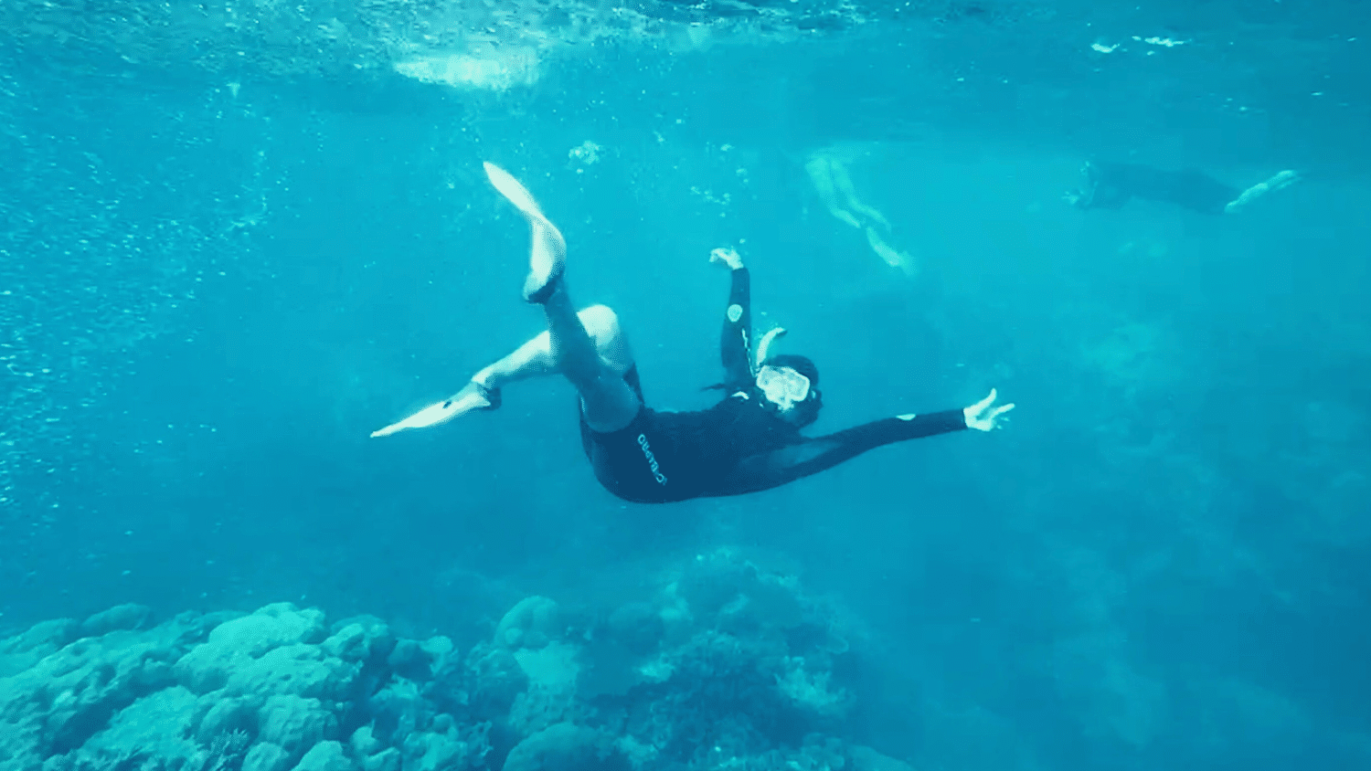 Annabelle Sharp poses underwater in a scuba mask, wet suit and flippers. She is in a body of bright blue water and a coral reef is a few feet below her, fading into the background.