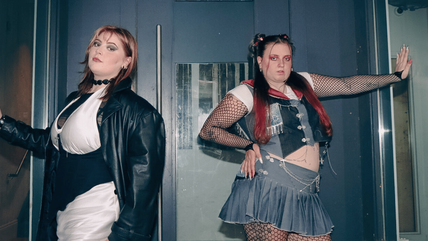 Two students pose in a doorway in front of blue double doors. On the left, a woman with shoulder-length hair wears a leather jacket, a black choker, and a white and black corseted dress. On the right of the image, the other student has a hand on her hip and her other pressed against a window. She has long hair with red ends, and wear a two piece denim, patchwork set with fishnet sleeves and tights.