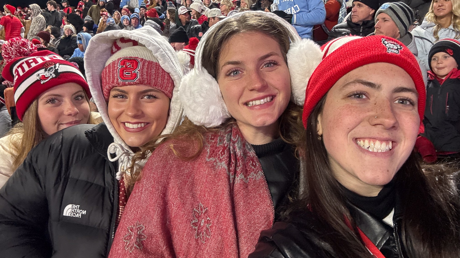 Penny Kudlak (far right) and three other people in winter clothing and NC State hats and earmuffs smile for a selfie at a football game, with a crowd of bundled-up people in the background.