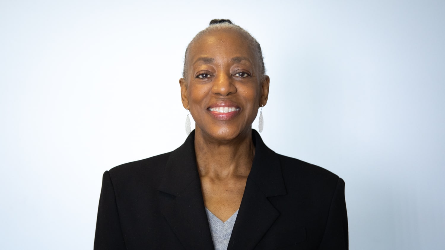 Sarah Wilmot smiles for a headshot. She has her hair in a slicked back bun, dangling earrings, and a black blazer on.