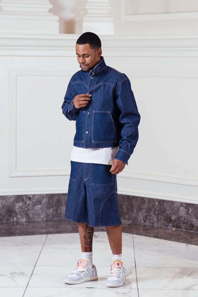 A model stands indoors wearing a dark blue denim jacket and matching knee-length denim shorts over a white shirt. They also wear white sneakers with pink accents and have visible tattoos on their left shin.